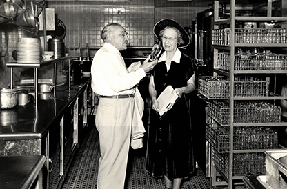 Man examining a glass while woman with a clipboard looks on.