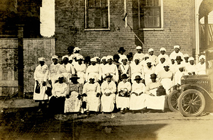 Group photo of African American midwives seated and standing outside of building.