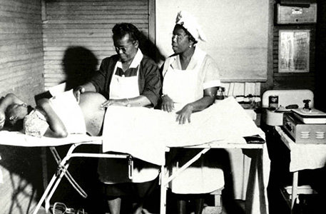 Black and white image of African American midwives examining the belly of a pregnant patient laying on an examination table.